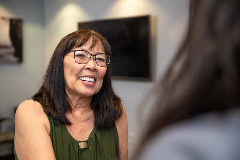 Middle-aged Asian woman smiling with all-on-4 dental implants talking with Silegy OMFS receptionist.